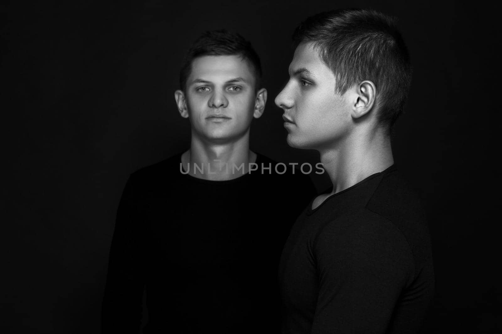 Two attractive positive young twin brothers standing dressed in black sweaters on black background. Black and white photography