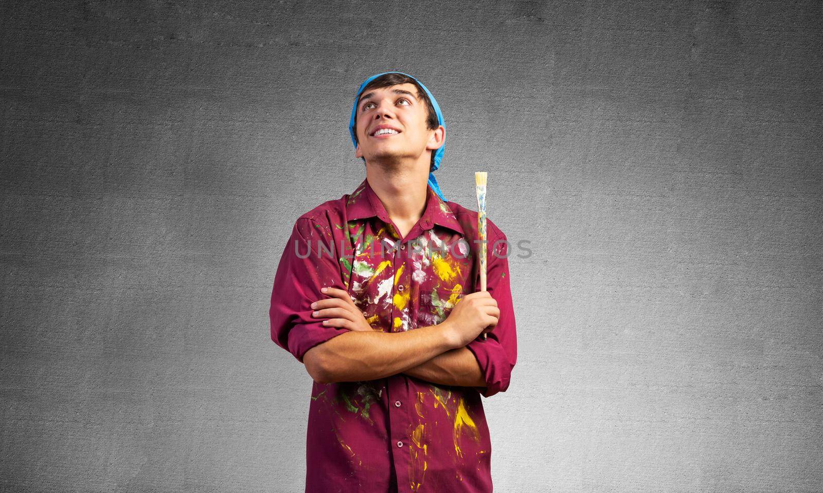 Smiling young painter artist with folded arms. Portrait of happy decorator looking upwards on gray wall background. Artistic occupation and creative profession. Art school student posing.