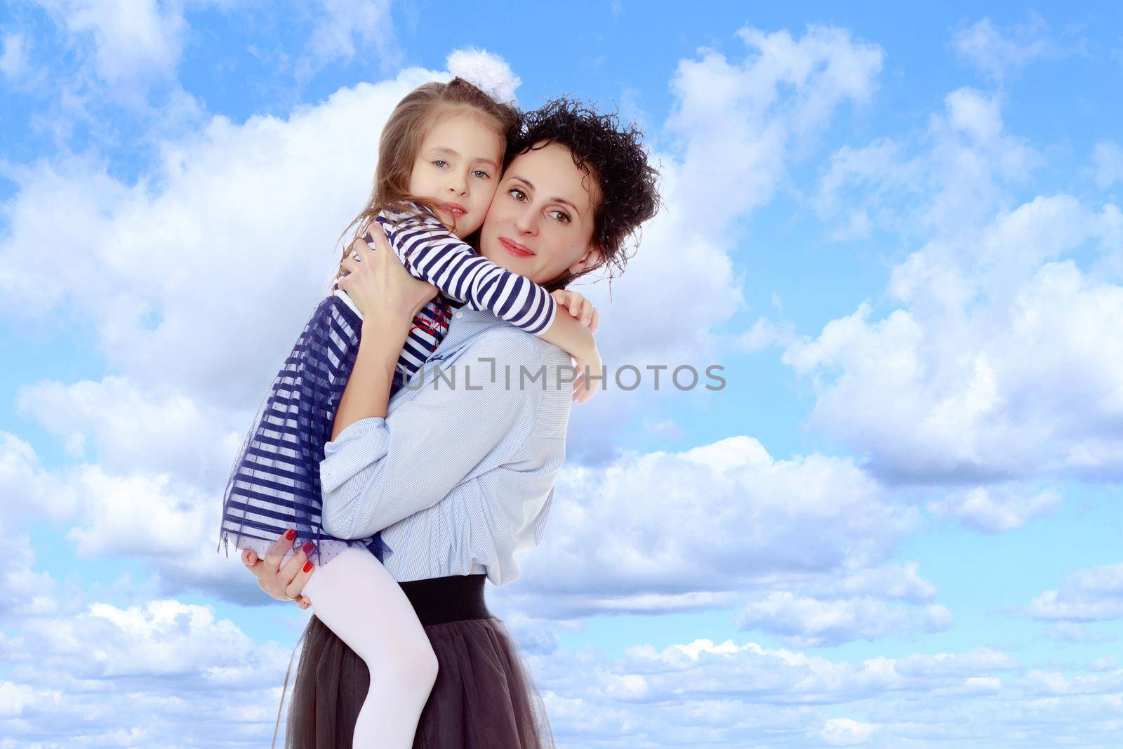 Happy young beautiful mother,lifted his little beloved daughter .And the girl embraced her mother's neck and pressed my face to her cheek.On the background of summer blue sky and fluffy clouds.