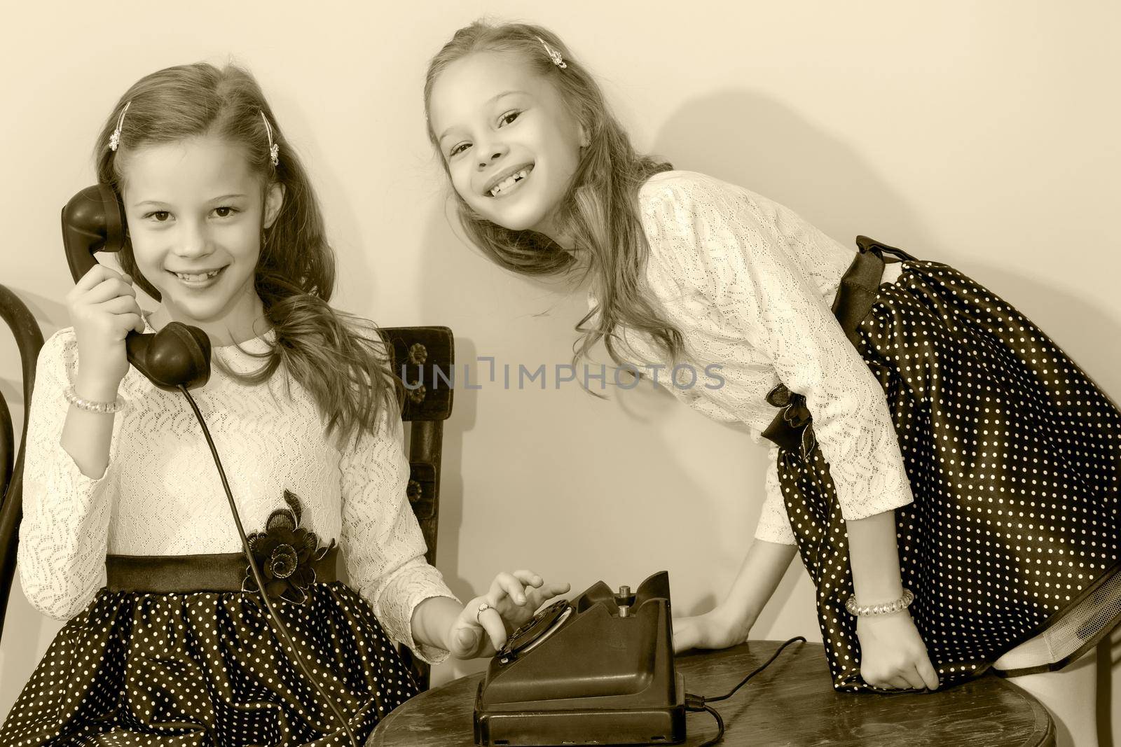 Two elegant girls sisters in beautiful dresses, talking on old phone. Black-and-white photo. Retro style.