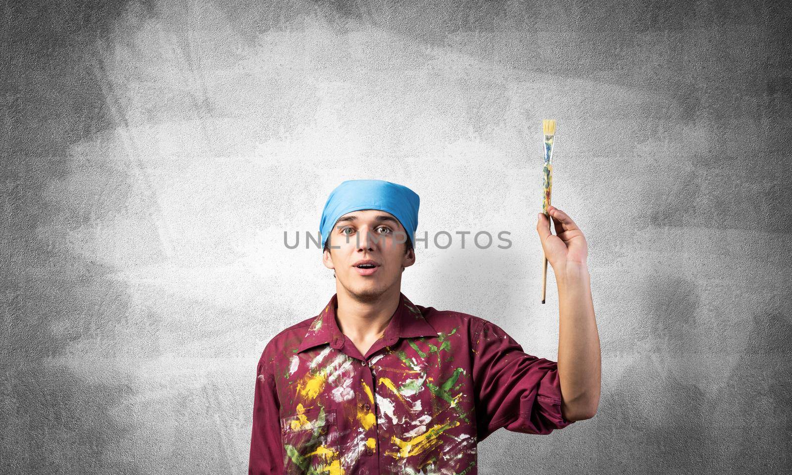 Young painter artist pointing upwards. Man holds paintbrush over his head. Portrait of caucasian painter on gray wall background. Creative hobby and artistic occupation. Art school student posing.