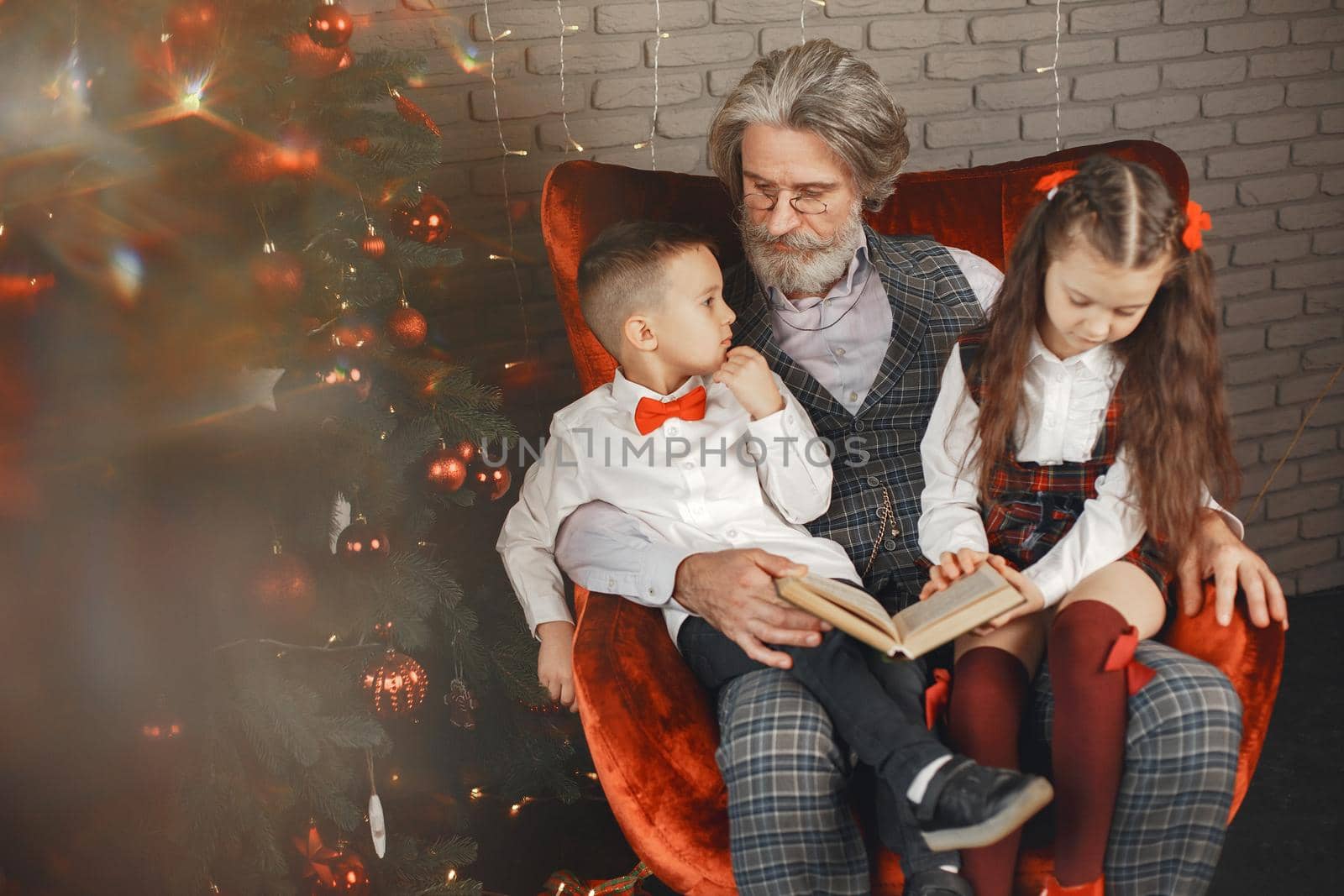 Grandfather wearing glasses, reading a book to small granddaughters twins in a room decorated for Christmas on the background of a Christmas tree. Christmas holiday concept. contrast photography