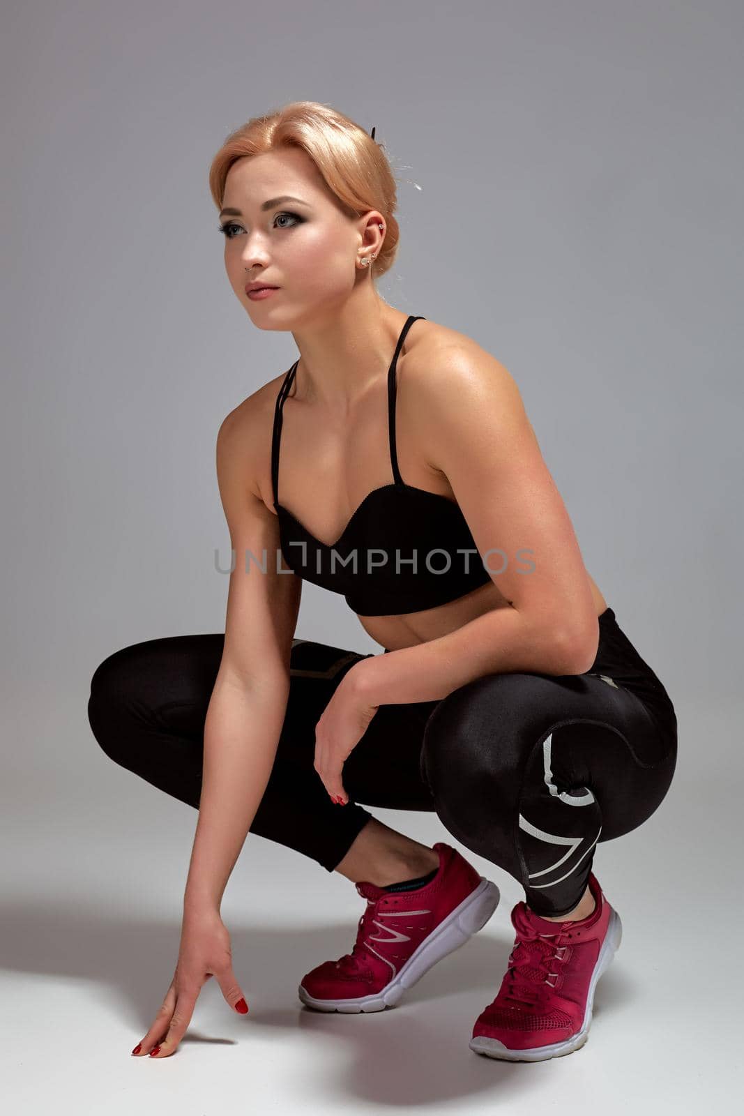 Happy cute young sportswoman sitting over gray background. Studio shot. Young woman wears black leggings and sports top