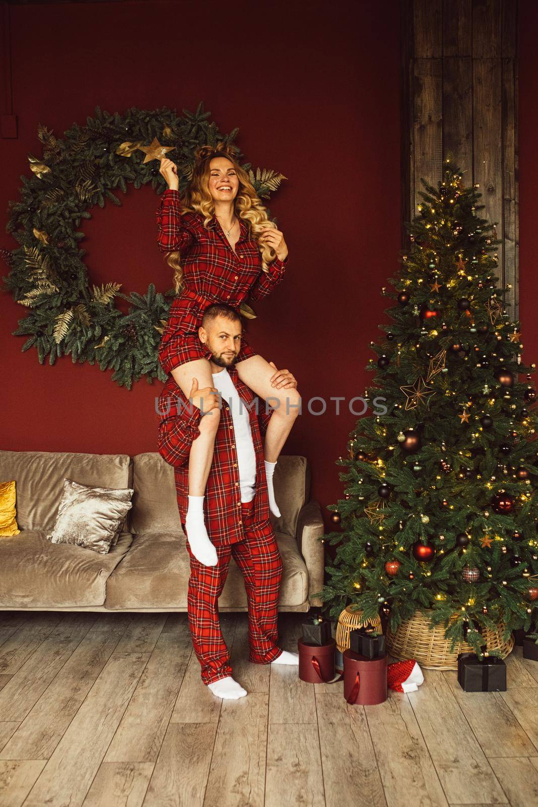 Pretty young lady sitting on her husband shoulders and decorating Christmas tree by StudioLucky