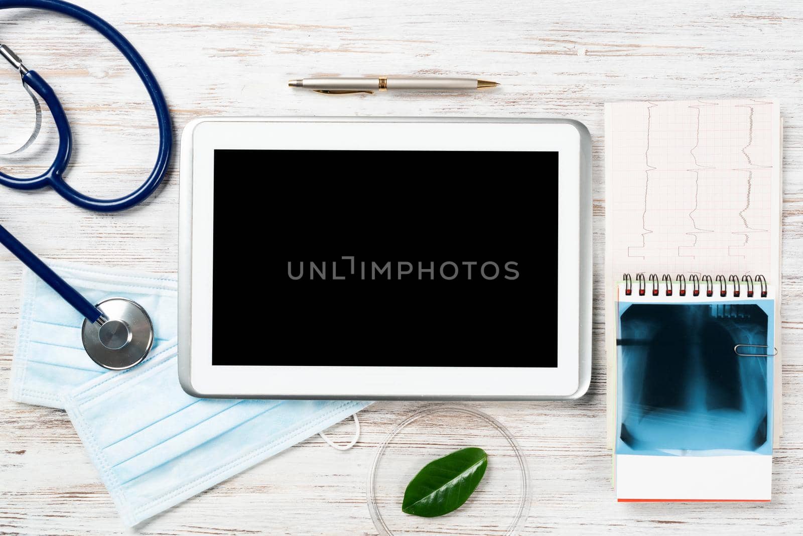 Flat lay workplace of doctor in modern clinic. Tablet computer with blank screen, x-ray image, stethoscope and cardiogram lies on wooden desk. Professional medical consultation and treatment