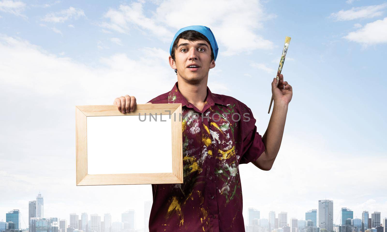 Young handsome painter artist holding paintbrush and blank canvas in frame. Portrait of happy painter on business district background. Creative hobby and artistic occupation. Art school student posing