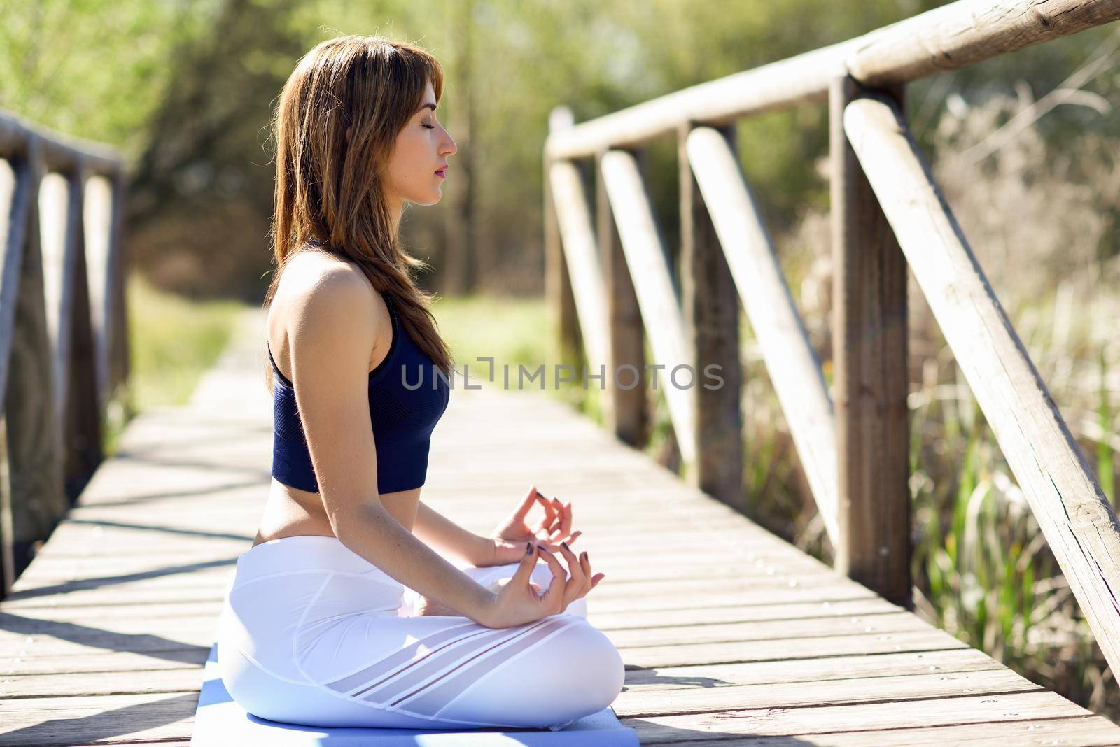 Young woman doing yoga in nature. Female wearing sport clothes in lotus figure. Girl training in a wooden bridge.