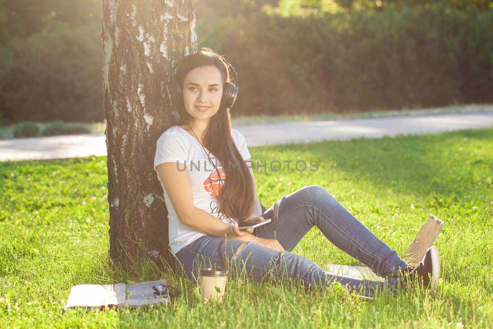 Woman relax with headphones listening to music sitting on grass in park. Young woman enjoys music and loneliness. Sun flare