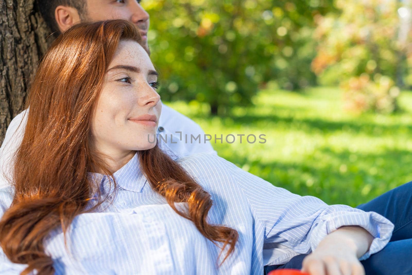 Young couple relaxing under tree in summer park on sunny day. Happy couple in love spend time outdoors together. Handsome man and pretty redhead girl enjoying each other. Romantic relationships.
