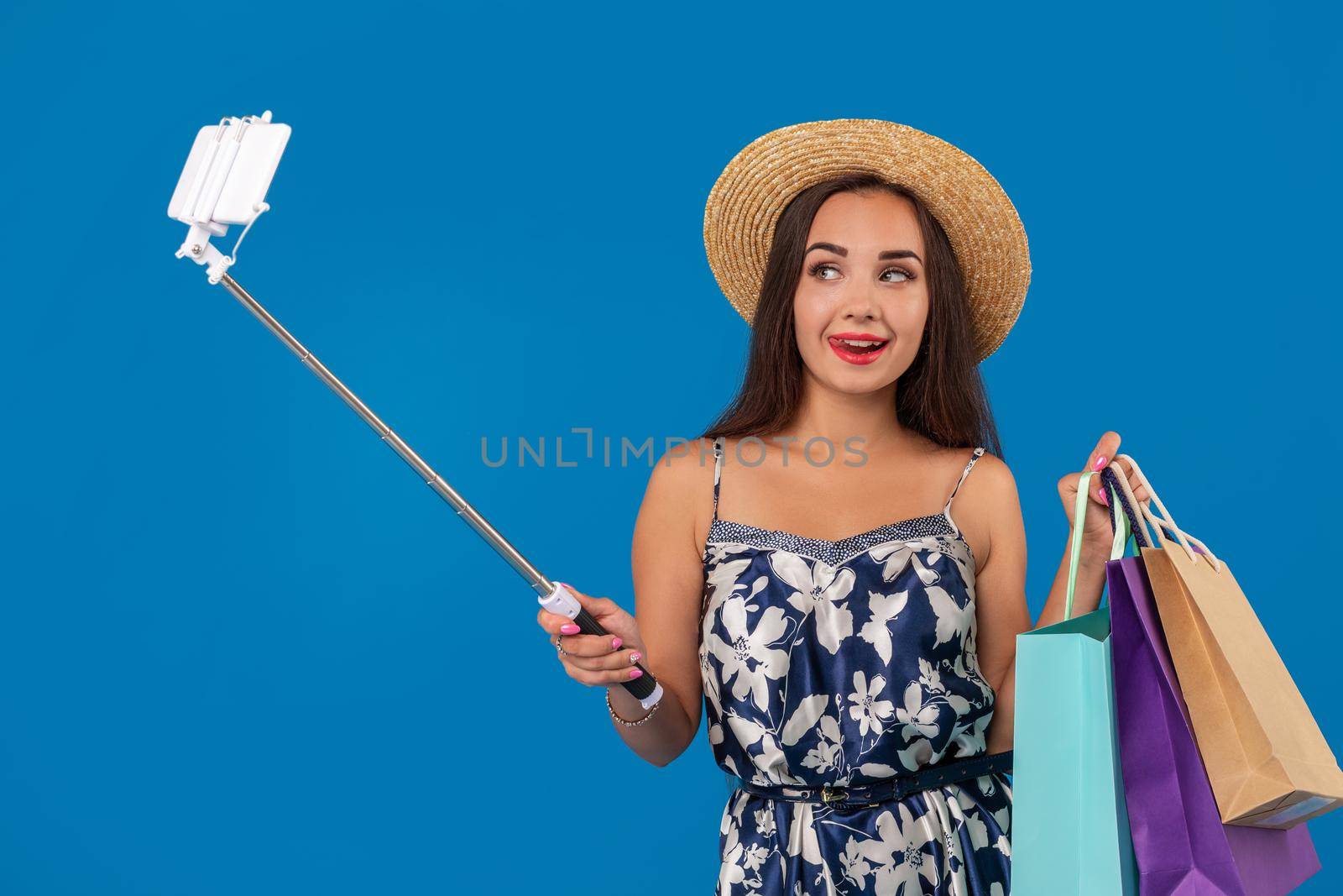 Trendy cheerful woman in casual clothes with shopping bags using selfie stick to take a self portrait on blue studio background with copy space. Emotions. Emotional woman