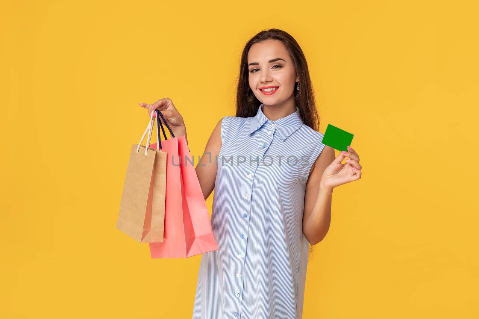 Trendy woman 20s in dress with long brown hair smiling while holding different shopping packages and card in hands isolated over yellow background. Shopping. Sale