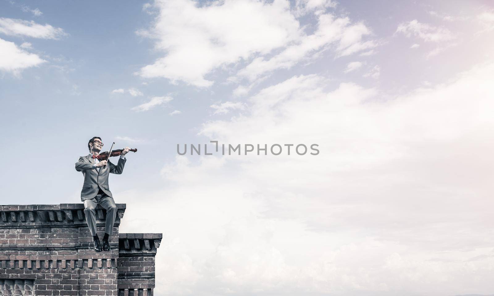 Young man wearing suit and glasses sitting on roof and playing violin