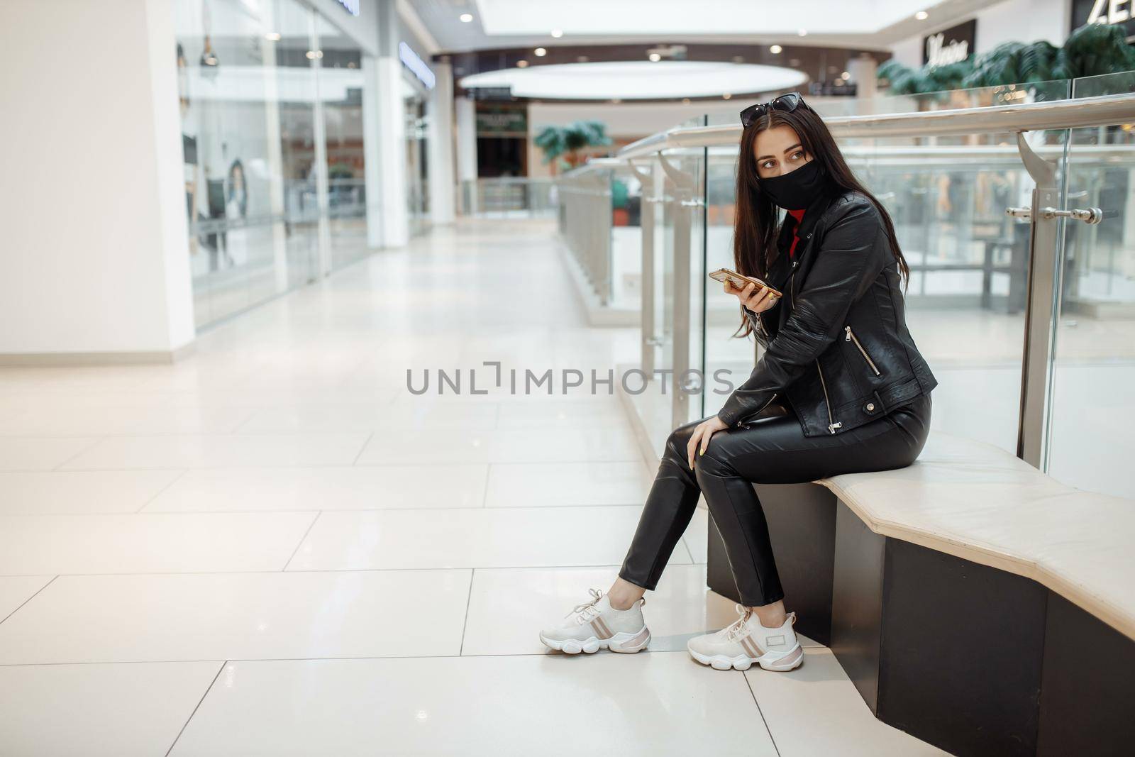 Girl with medical black mask and mobile phone in a shopping center. Coronavirus pandemic. A woman with a mask is standing in a shopping center. A girl in a protective mask is shopping at the mall.