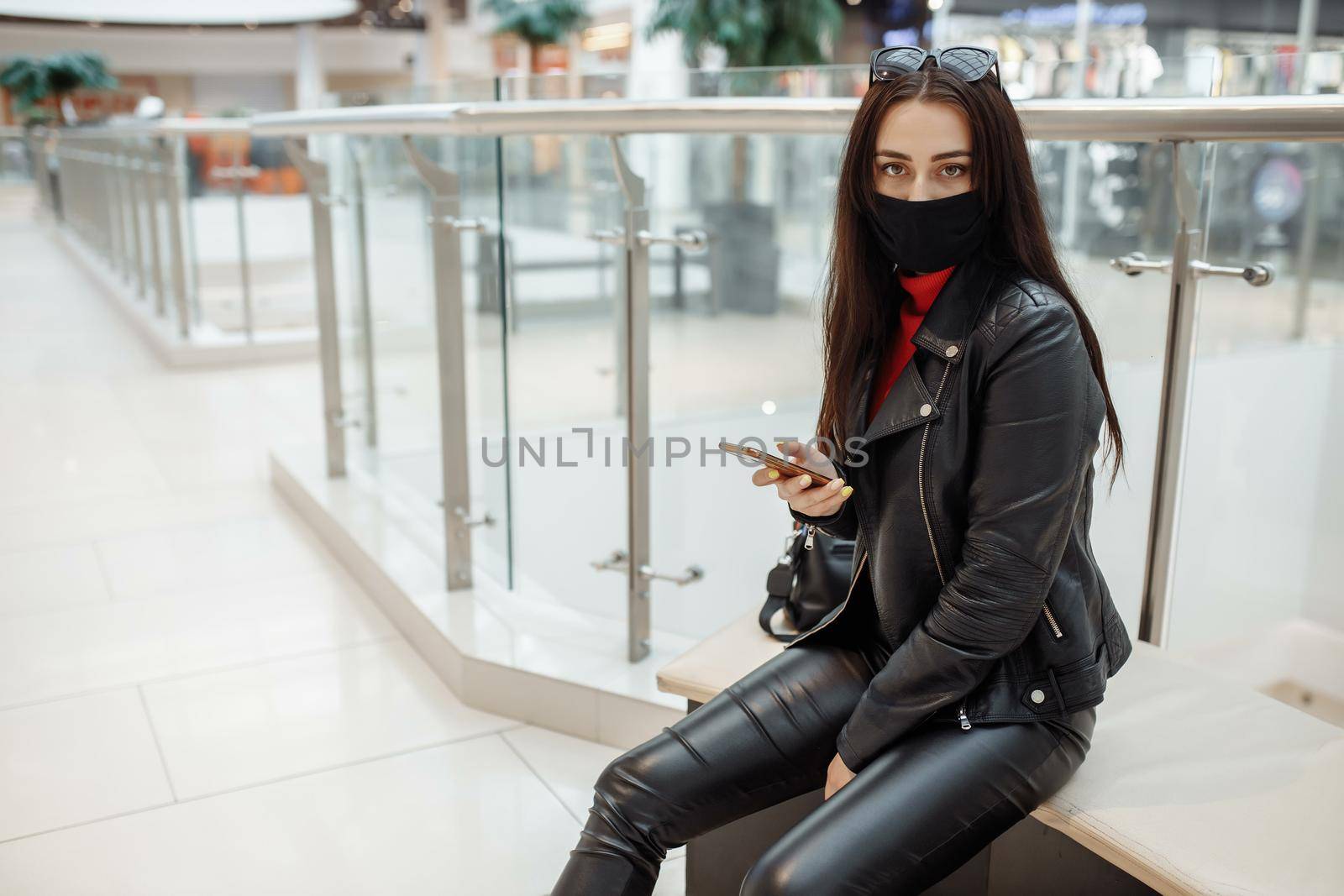 Girl with medical black mask and mobile phone in a shopping center. Coronavirus pandemic. A woman with a mask is standing in a shopping center. A girl in a protective mask is shopping at the mall by UcheaD