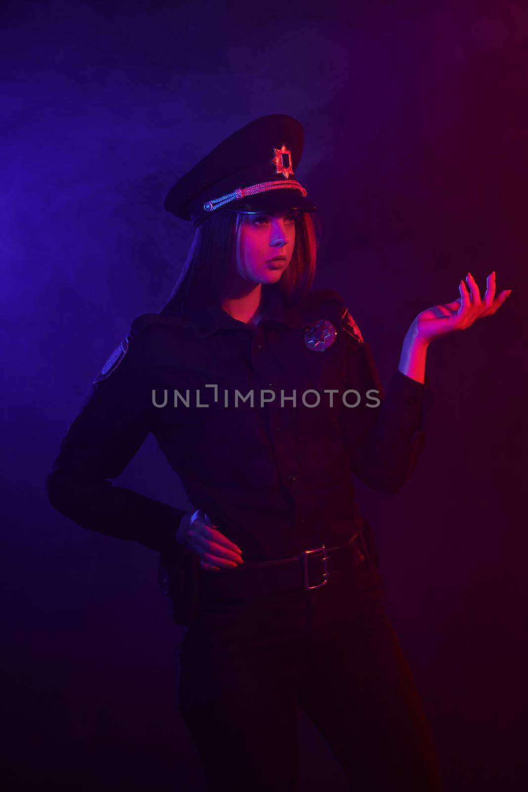 Pretty redheaded maiden police officer in a uniform and a cap, with bright make-up is looking away and posing against a black background with red and blue backlighting. Defender of citizens is ready to enforce a law and stop a crime.