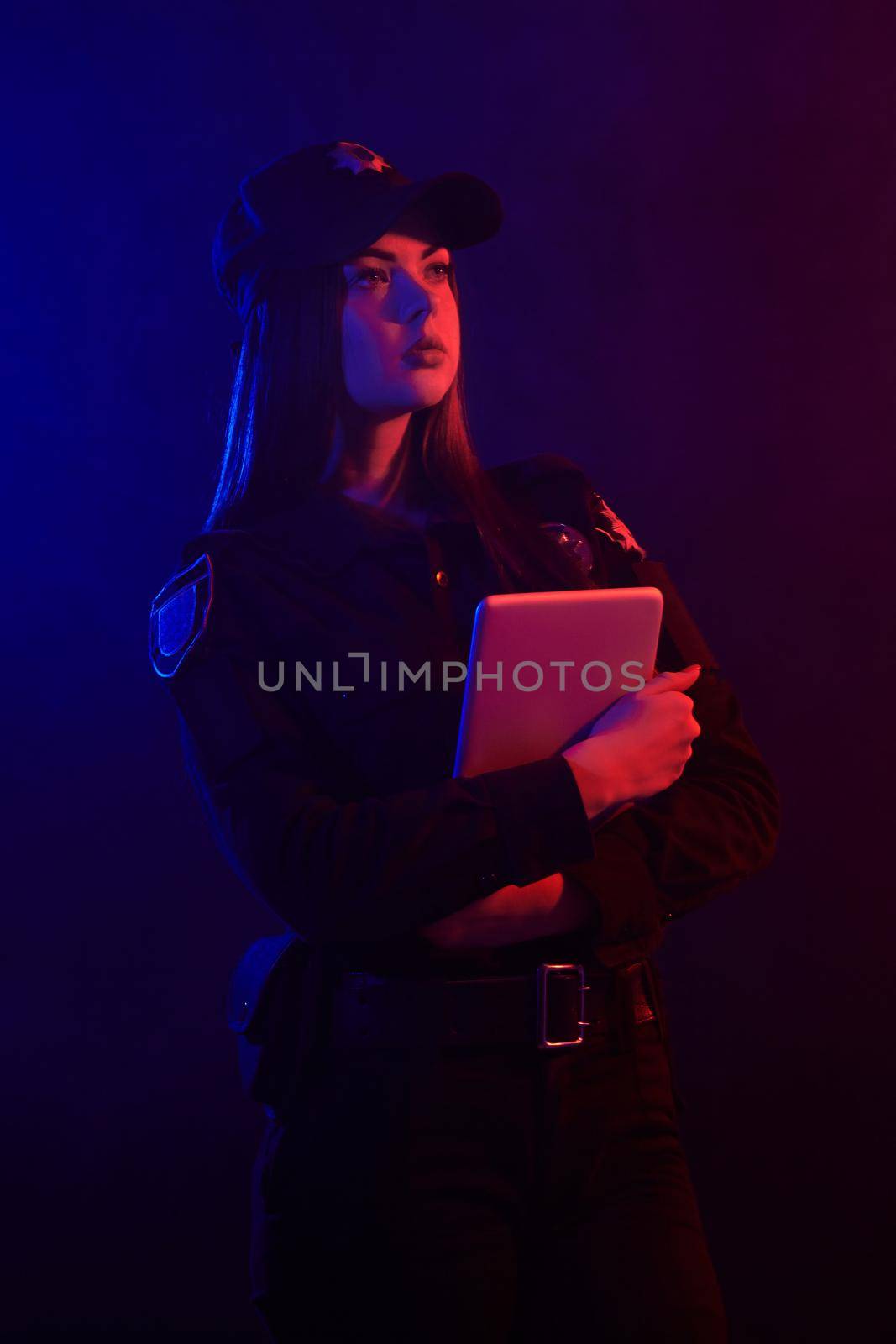 Beautiful young female police officer in a uniform and a cap, with bright make-up is holding her tablet and posing sideways against a black background with red and blue backlighting. Defender of citizens is ready to enforce a law and stop a crime.