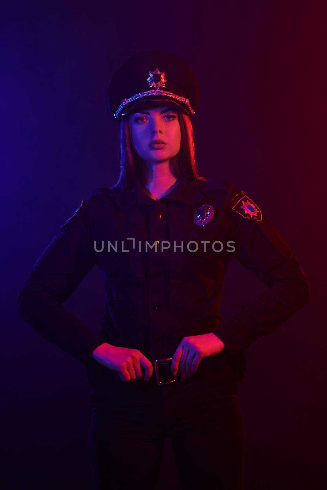 Young redheaded female police officer in a uniform and a cap, with bright make-up looking at the camera and posing against a black background with red and blue backlighting. Defender of citizens is ready to enforce a law and stop a crime.