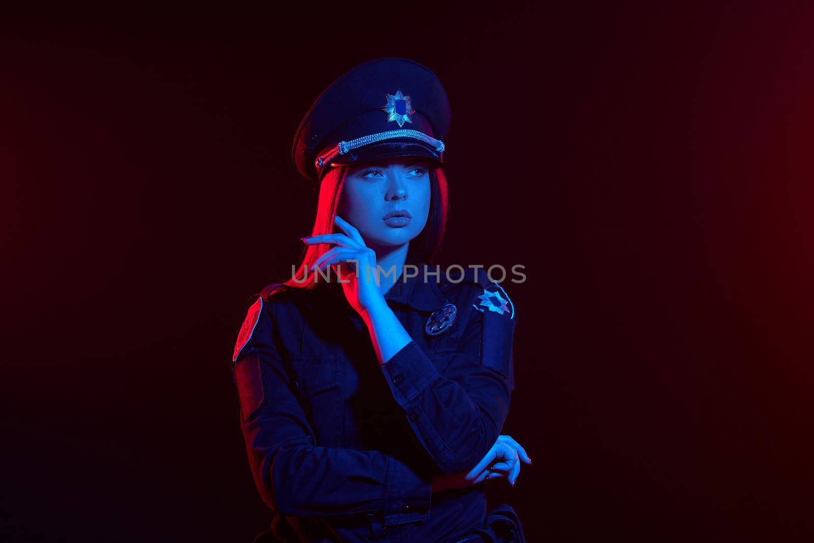 Close-up shot of an attractive redheaded woman police officer in a uniform and a cap looking away and posing with crossed hands against a black background with red and blue backlighting. Defender of citizens is ready to enforce a law and stop a crime.