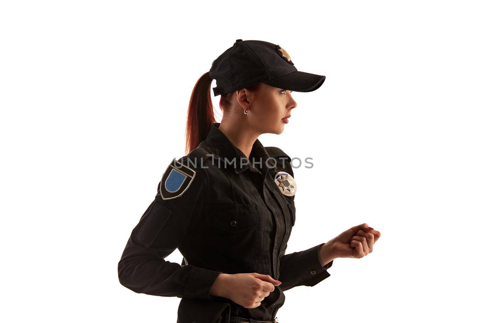 Close-up shot of a pretty redheaded policewoman in a black uniform and a cap preparing to fight, standing sideways isolated on white background. Defender of citizens is ready to enforce a law and stop a crime.