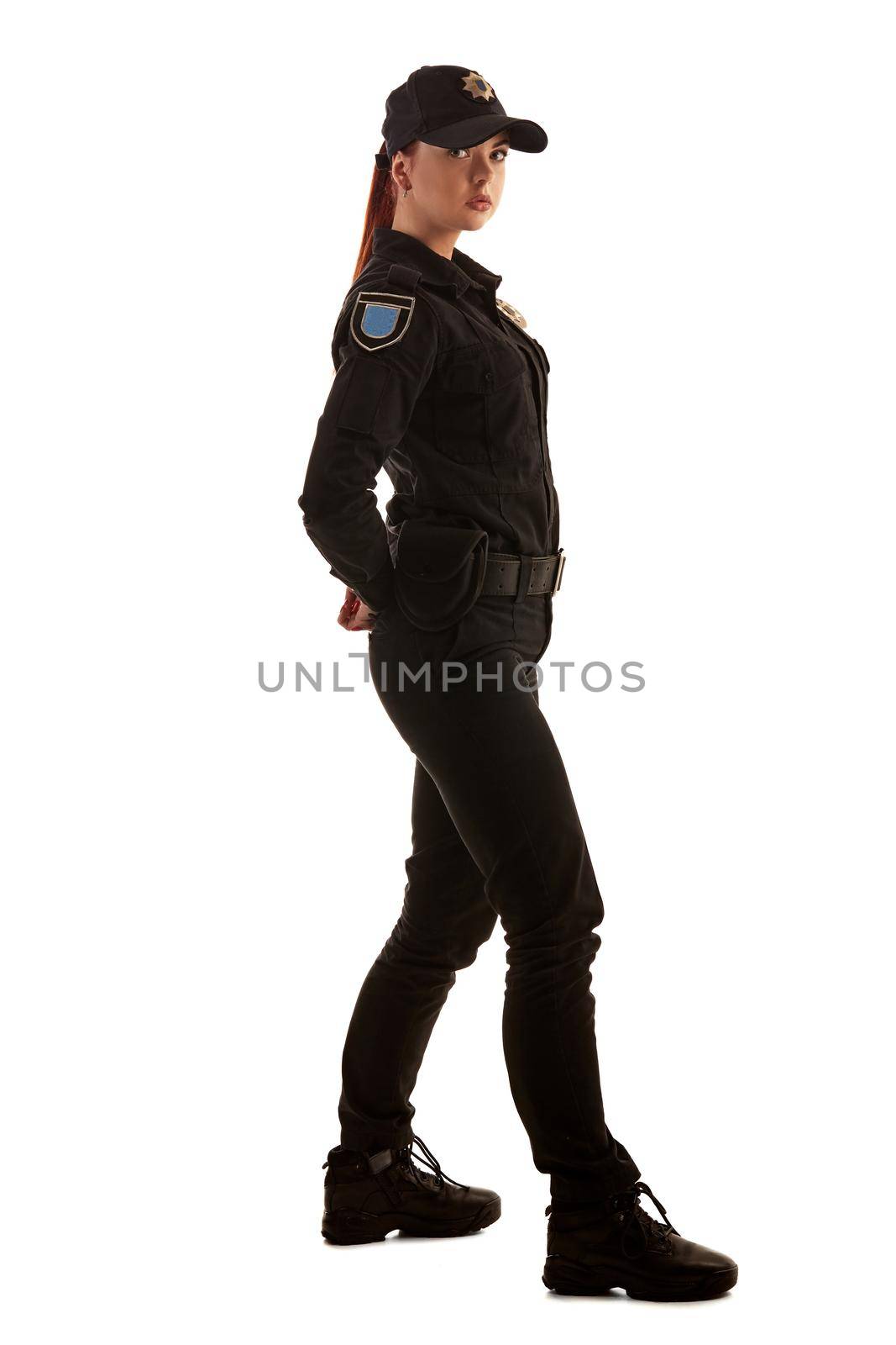 Full length shot of a beautiful redheaded woman police officer in a uniform and a cap posing sideways and looking at the camera, isolated on white background. Defender of citizens is ready to enforce a law and stop a crime.