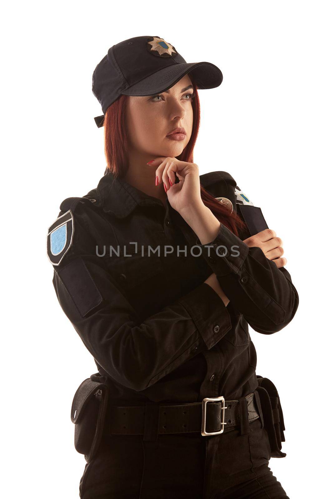 Beautiful ginger policewoman in a black uniform and a cap is looking away and posing standing sideways, isolated on white background. Defender of citizens is ready to enforce a law and stop a crime.