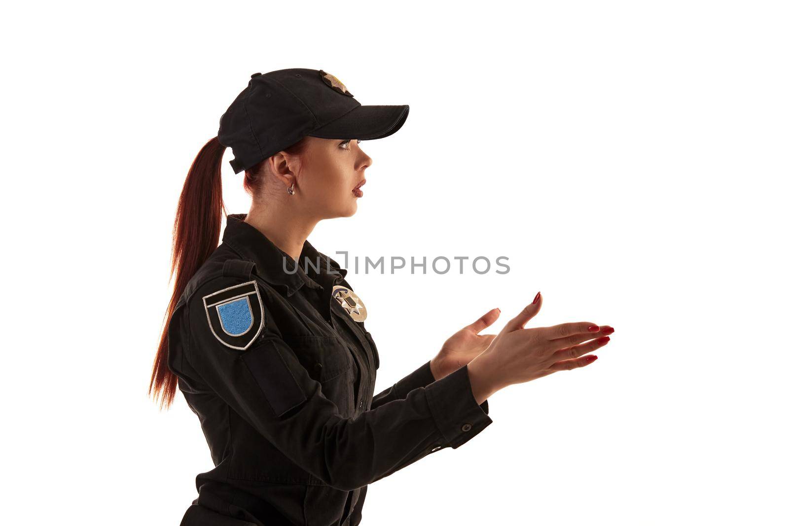 Close-up shot of a redheaded professional policewoman in a black uniform and a cap posing sideways and gesticulating, isolated on white background. Defender of citizens is ready to enforce a law and stop a crime.