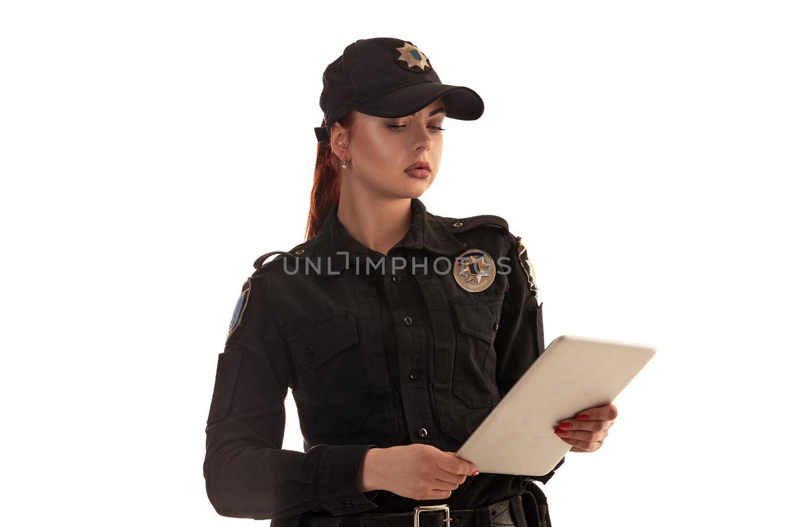 Close-up shot of a serious ginger policewoman in a black uniform and a cap holding a tablet in her arms and looking at it while posing isolated on white background. Defender of citizens is ready to enforce a law and stop a crime.