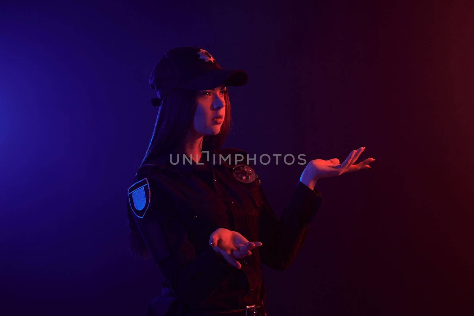 Beautiful redheaded lady police officer in a uniform and a cap, with bright make-up is gesticulating while posing sideways against a black background with red and blue backlighting. Defender of citizens is ready to enforce a law and stop a crime.