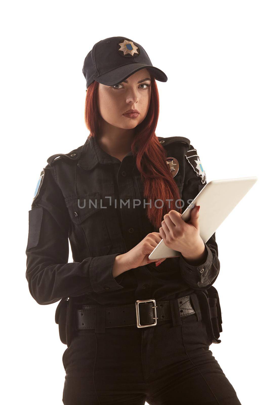 Sexy redheaded policewoman in a black uniform and a cap is holding a tablet in her arms and looking at the camera, isolated on white background. Defender of citizens is ready to enforce a law and stop a crime.
