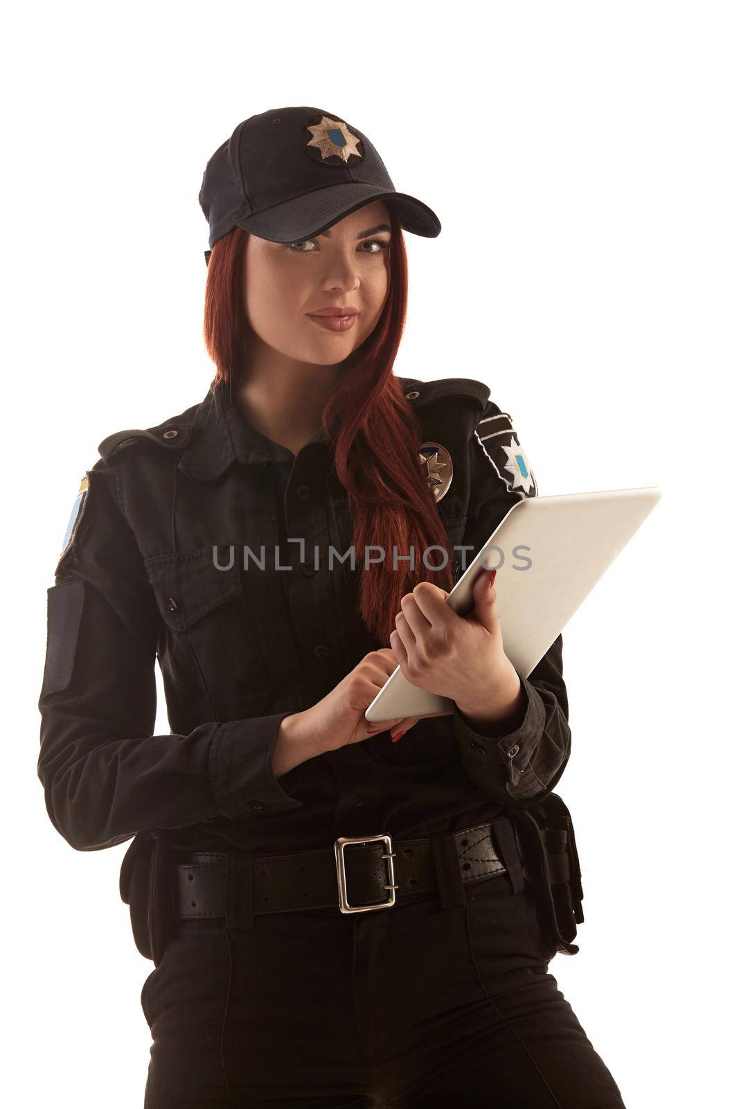 Cute ginger policewoman in a black uniform and a cap is holding a tablet in her arms, looking at the camera and smiling, isolated on white background. Defender of citizens is ready to enforce a law and stop a crime.