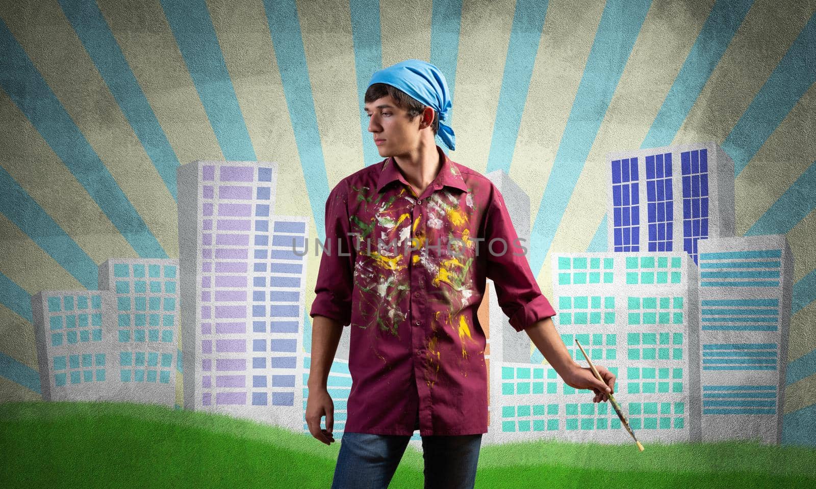Young male artist holding paintbrush. Painter in shirt and bandana standing on background colorful picture on wall. Modern cityscape with high skyscrapers artwork. Creative hobby and profession