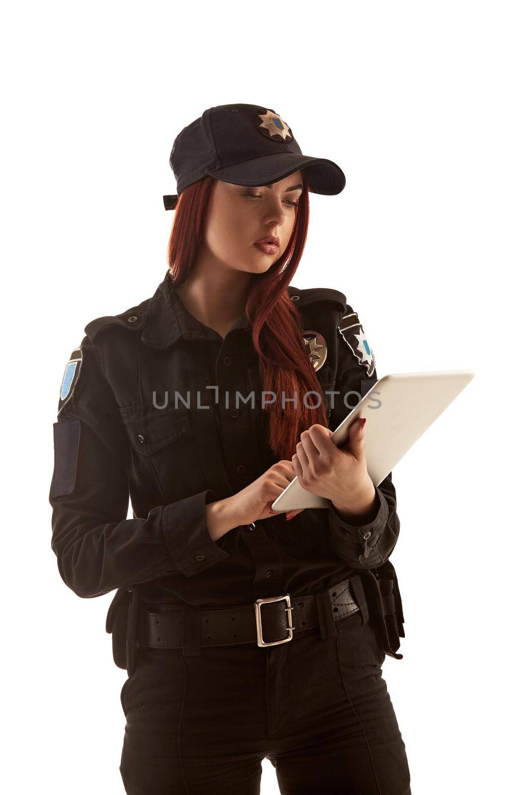 Lovely ginger policewoman in a black uniform and a cap is holding a tablet in her arms and typing something on it, isolated on white background. Defender of citizens is ready to enforce a law and stop a crime.