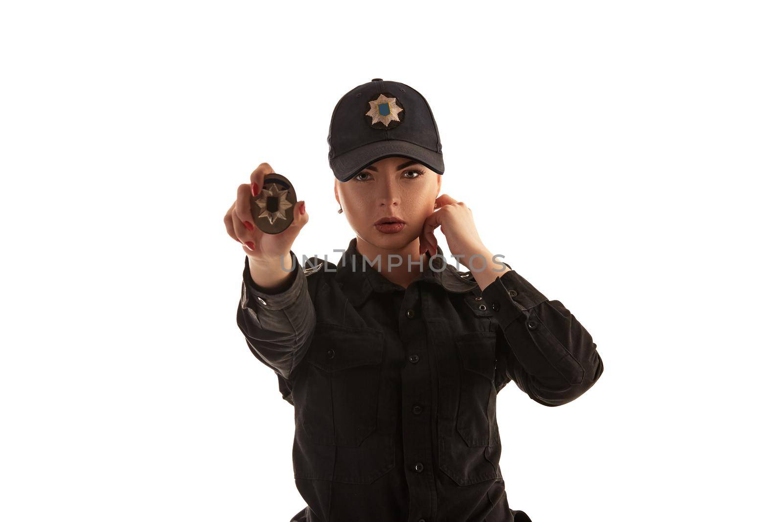 Close-up shot of a strong redheaded policewoman in a black uniform and a cap showing her badge and looking at the camera, isolated on white background. Defender of citizens is ready to enforce a law and stop a crime.