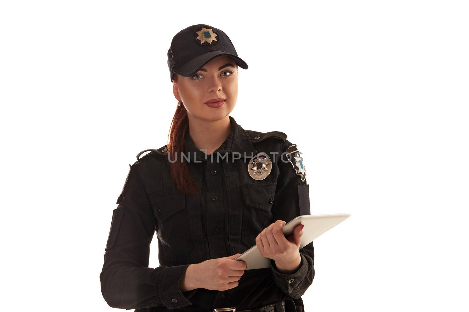 Close-up shot of a pretty redheaded policewoman in a black uniform and a cap holding a tablet in her arms. She is looking at the camera and smiling while posing isolated on white background. Defender of citizens is ready to enforce a law and stop a crime.