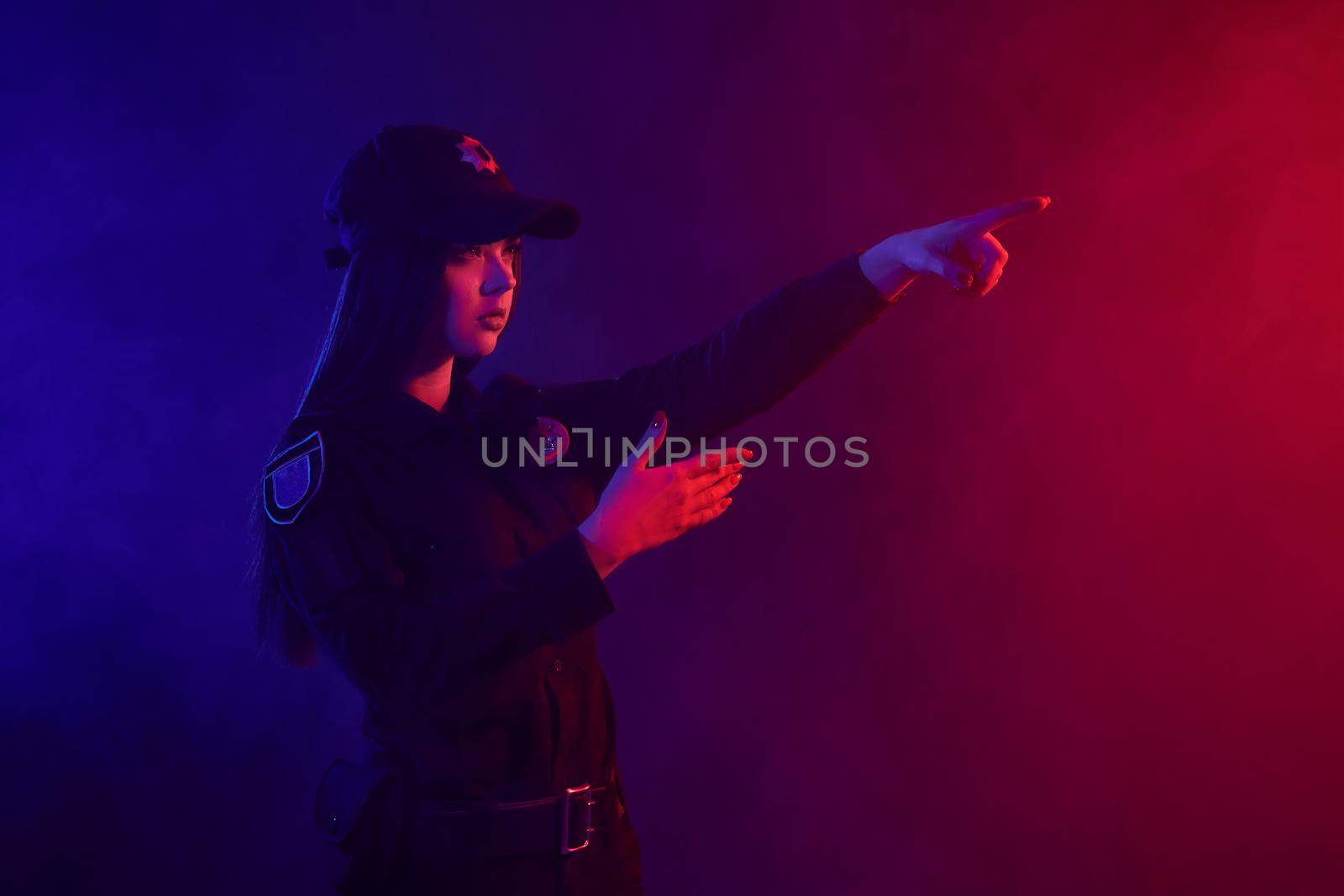 Charming redheaded female traffic controller in a uniform and a cap is regulating traffic with signals posing sideways against a black background with red and blue backlighting. Defender of citizens is ready to enforce a law and stop a crime.