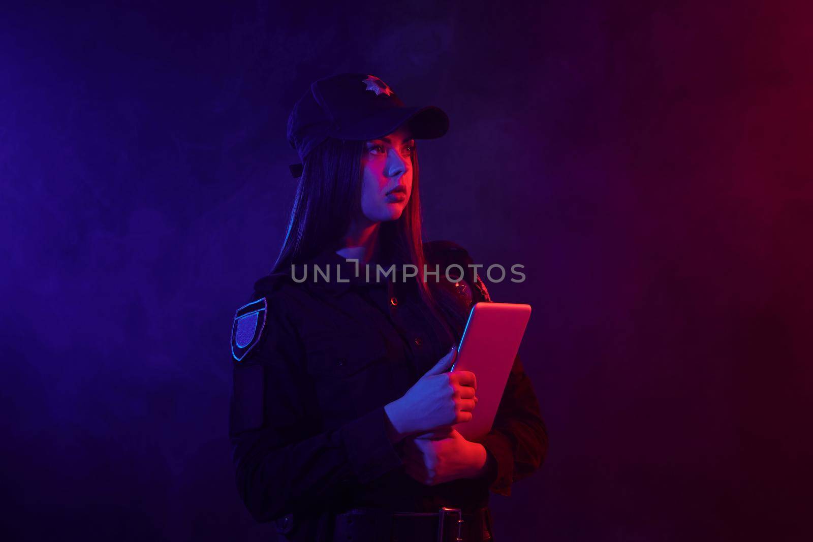 Beautiful redheaded woman police officer in a uniform and a cap, with bright make-up is holding her tablet and posing sideways against a black background with red and blue backlighting. Defender of citizens is ready to enforce a law and stop a crime.