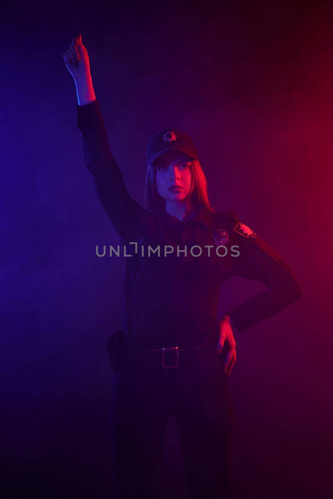Charming redheaded girl traffic controller in a uniform and a cap is looking at the camera and regulating traffic with signals against a black background with red and blue backlighting. Defender of citizens is ready to enforce a law and stop a crime.