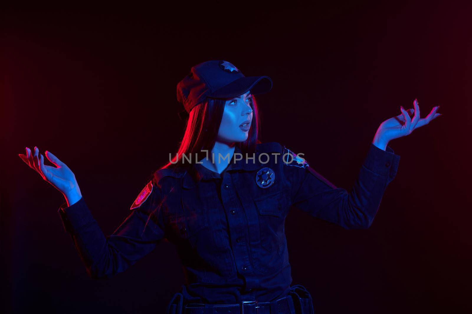 Close-up shot of an attractive redheaded lady police officer in a uniform and a cap looking away and gesticulating against a black background with red and blue backlighting. Defender of citizens is ready to enforce a law and stop a crime.