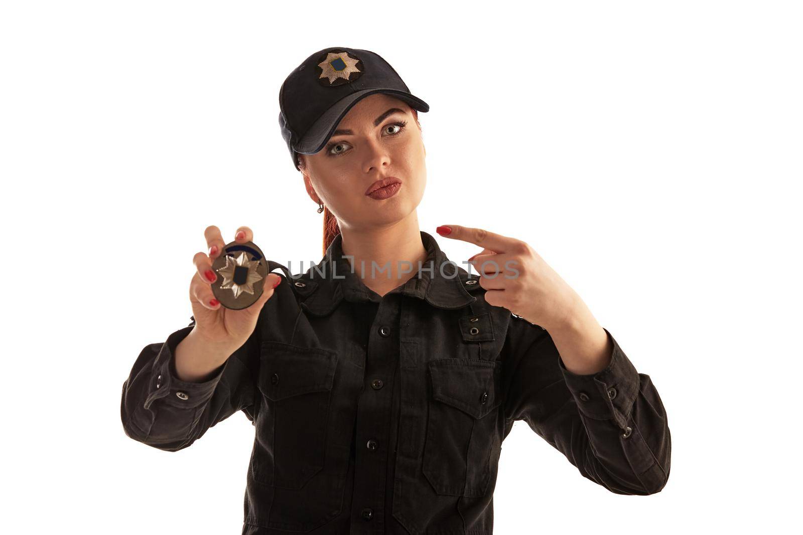 Close-up shot of a srious redheaded policewoman in a black uniform and a cap showing her badge, pointing on it and looking at the camera, isolated on white background. Defender of citizens is ready to enforce a law and stop a crime.