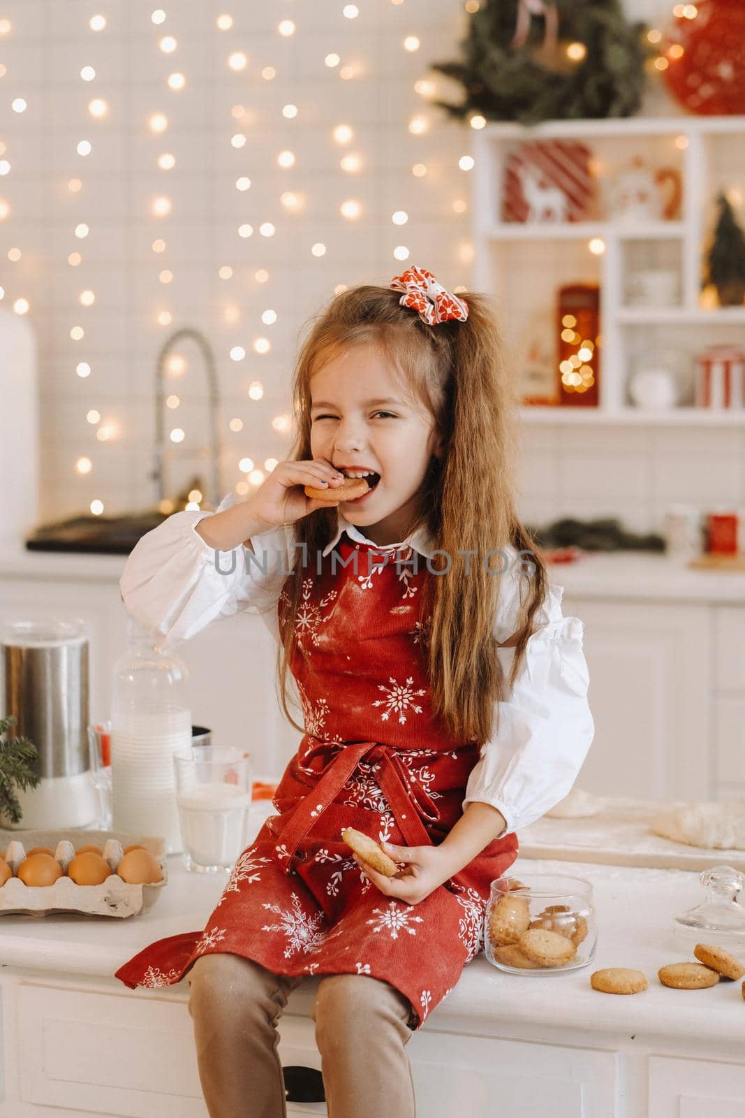 A happy little girl in the Christmas kitchen is sitting on the table with cookies in her hands and eating it.