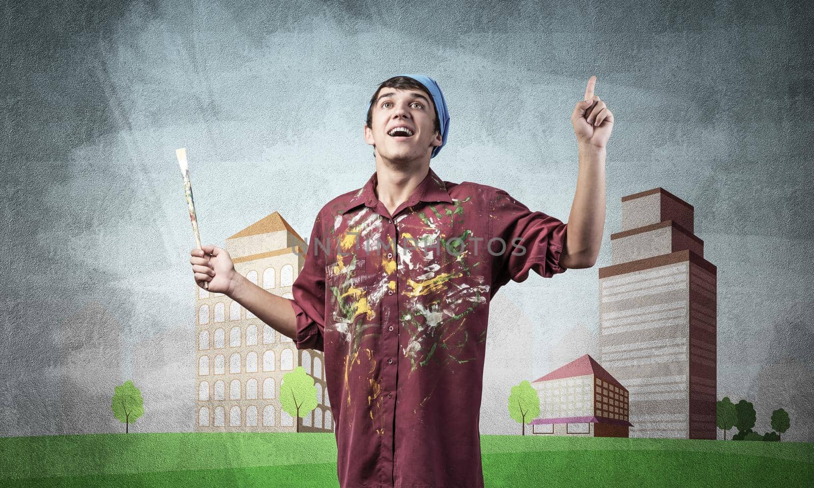 Young male artist holding paintbrush. Happy painter in shirt and bandana standing on background colorful picture on wall. Modern cityscape with high skyscrapers artwork. Creative hobby and profession