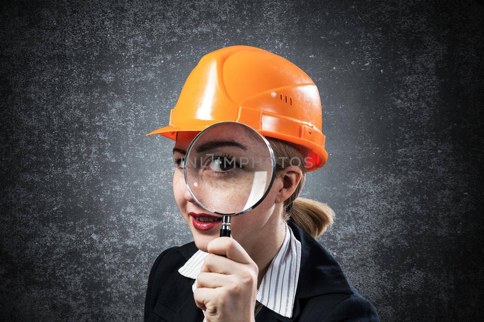 Building inspector looking through magnifying glass. Young construction specialist in safety helmet with magnifier on grey wall background. Consultancy and expertise. Professional valuation services
