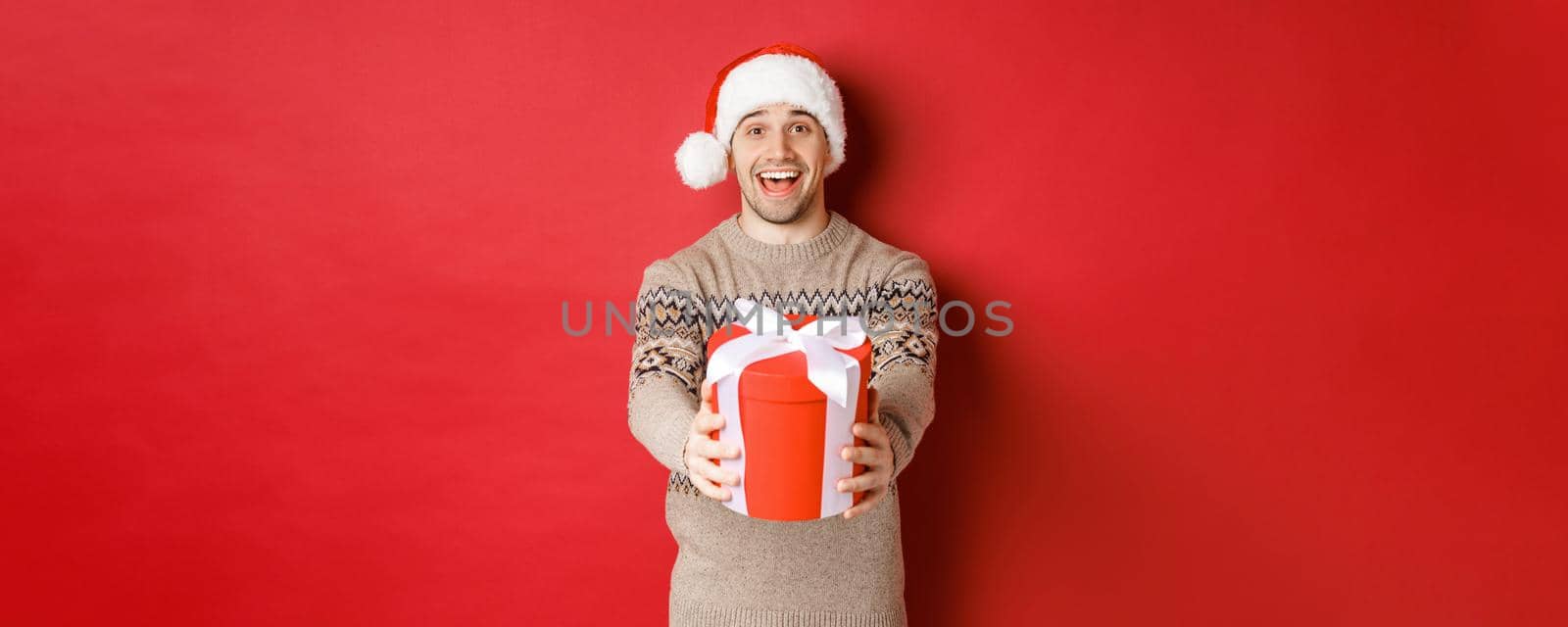 Image of happy man saying merry christmas and giving you a gift in box, smiling cheerful, celebrating winter holidays in santa hat and xmas sweater, standing over red background.