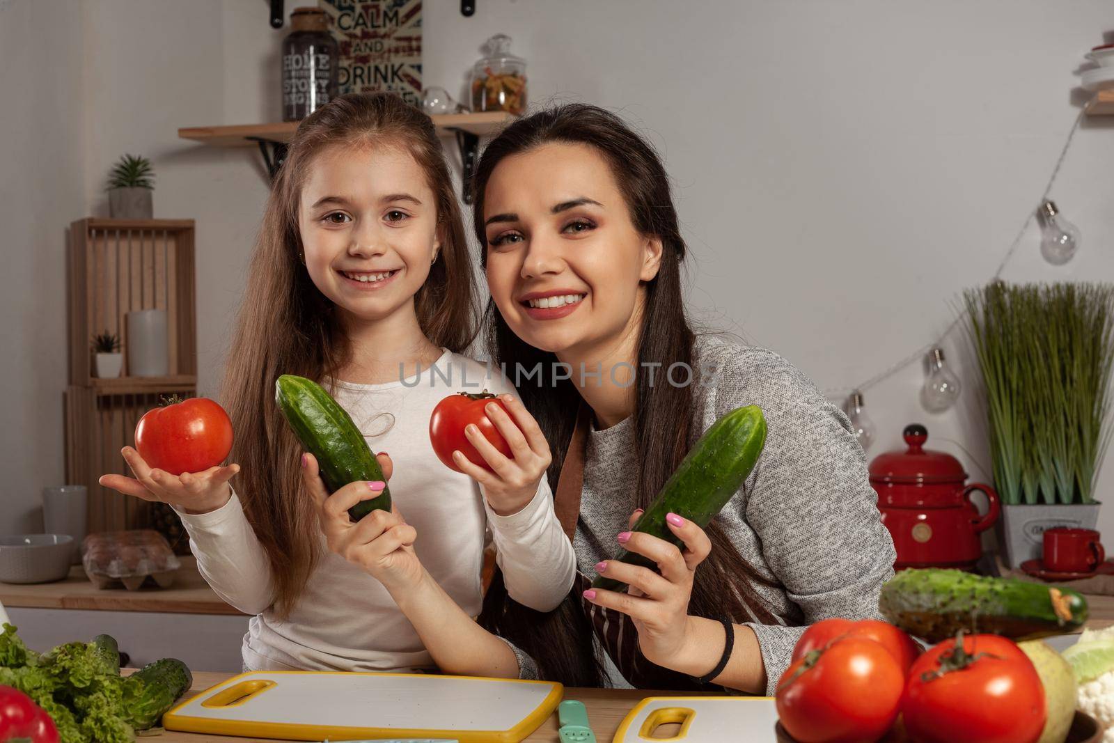 Happy loving family are cooking together. Delightful mum and her child are posing with tomato and cucumber and smiling at the kitchen, against a white wall with shelves and bulbs on it. Homemade food and little helper.