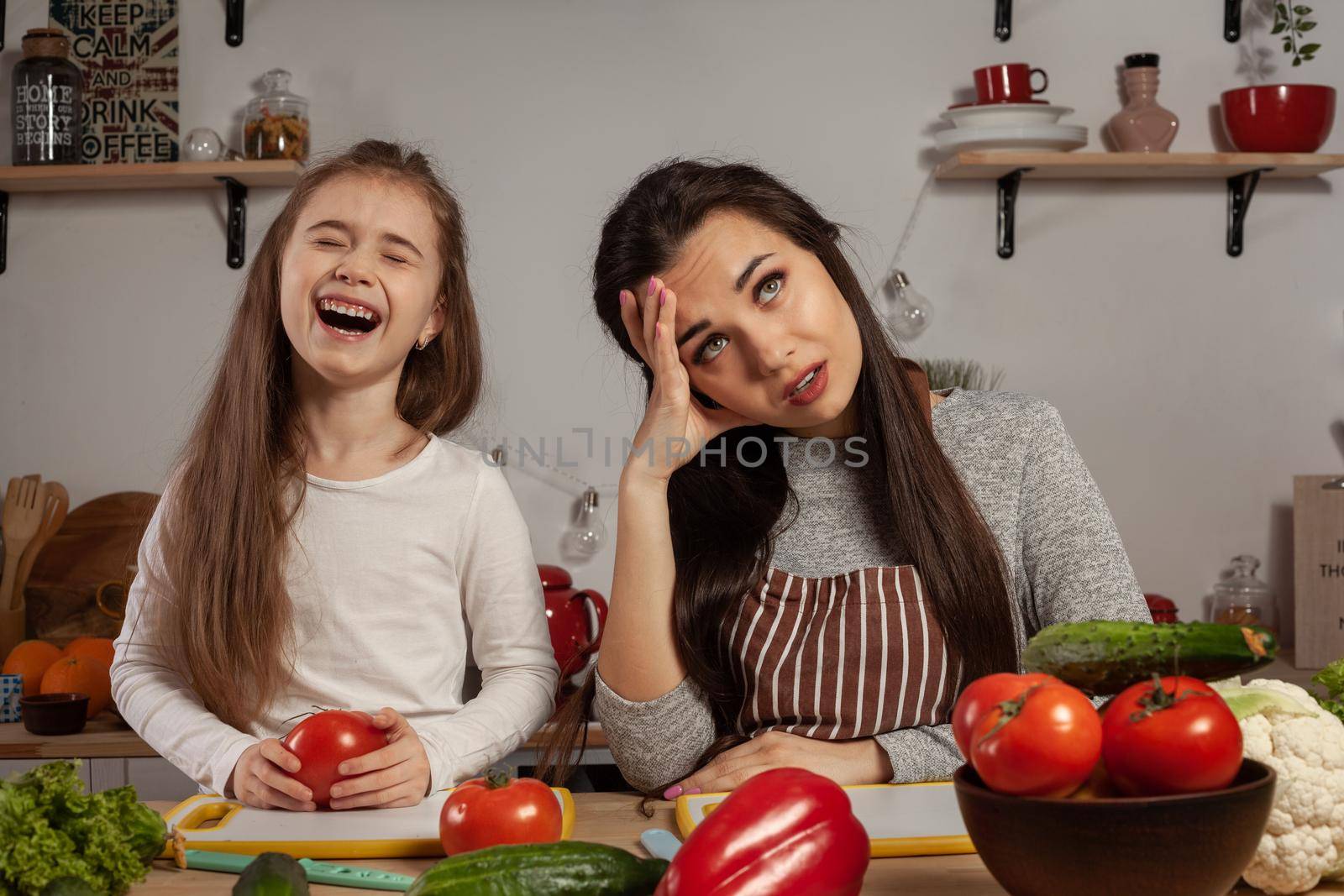 Happy loving family are cooking together. Lovely mommy and her little girl are making a vegetable salad and tired of each other at the kitchen, against a white wall with shelves and bulbs on it. Homemade food and little helper.