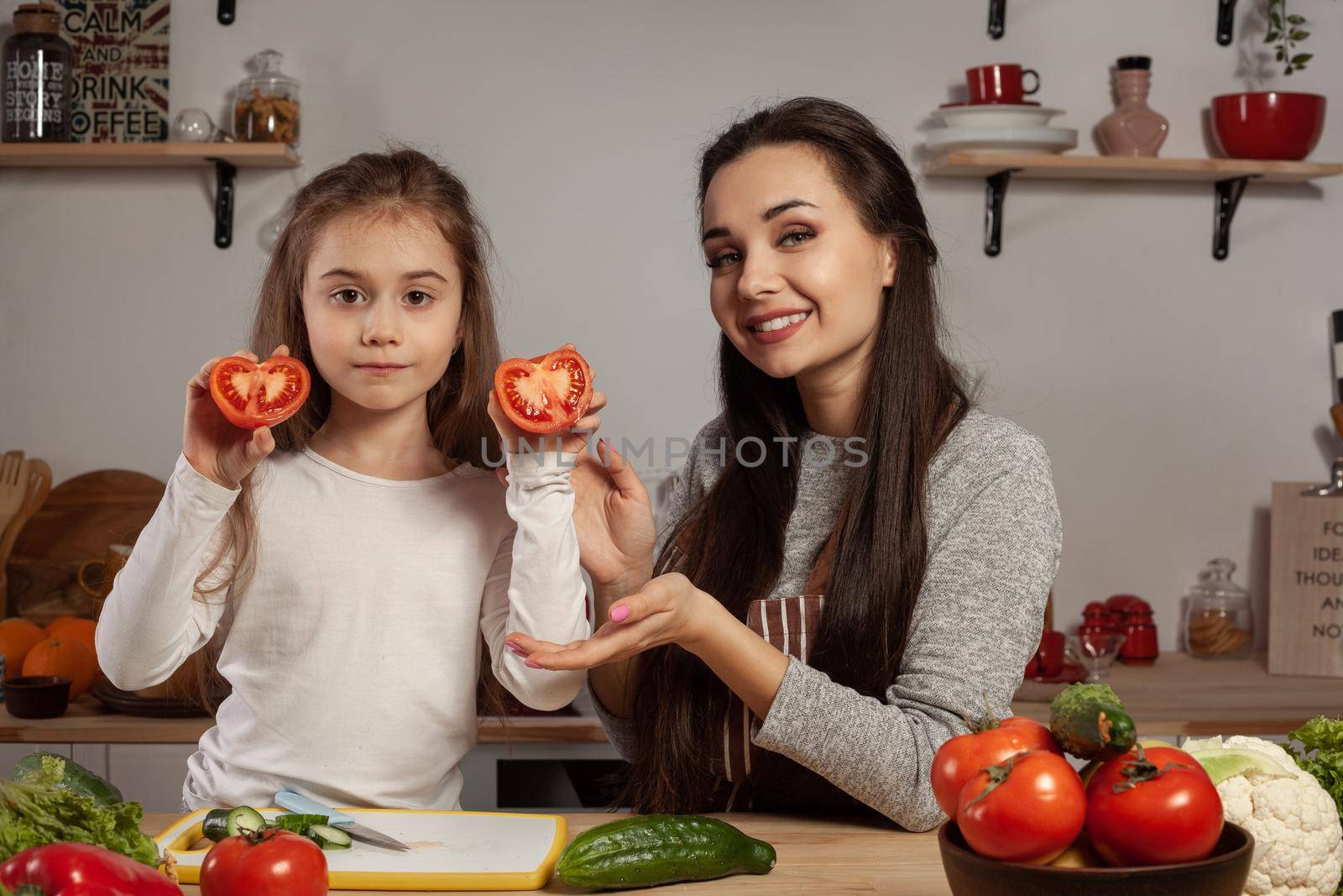 Happy loving family are cooking together. Pretty mom and her kid are making a vegetable salad and posing with tomato at the kitchen, against a white wall with shelves and bulbs on it. Homemade food and little helper.