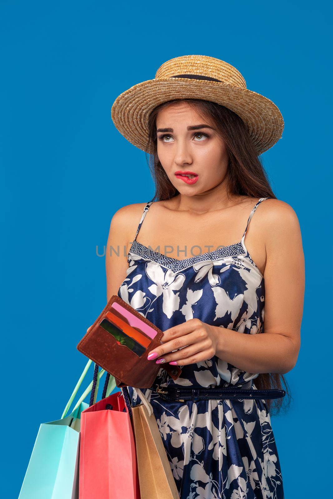 Portrait of unhappy young woman looking in her wallet in shopping center, spent too much, not enough cash, lost money, broke. Studio shot on blue background. Copy space