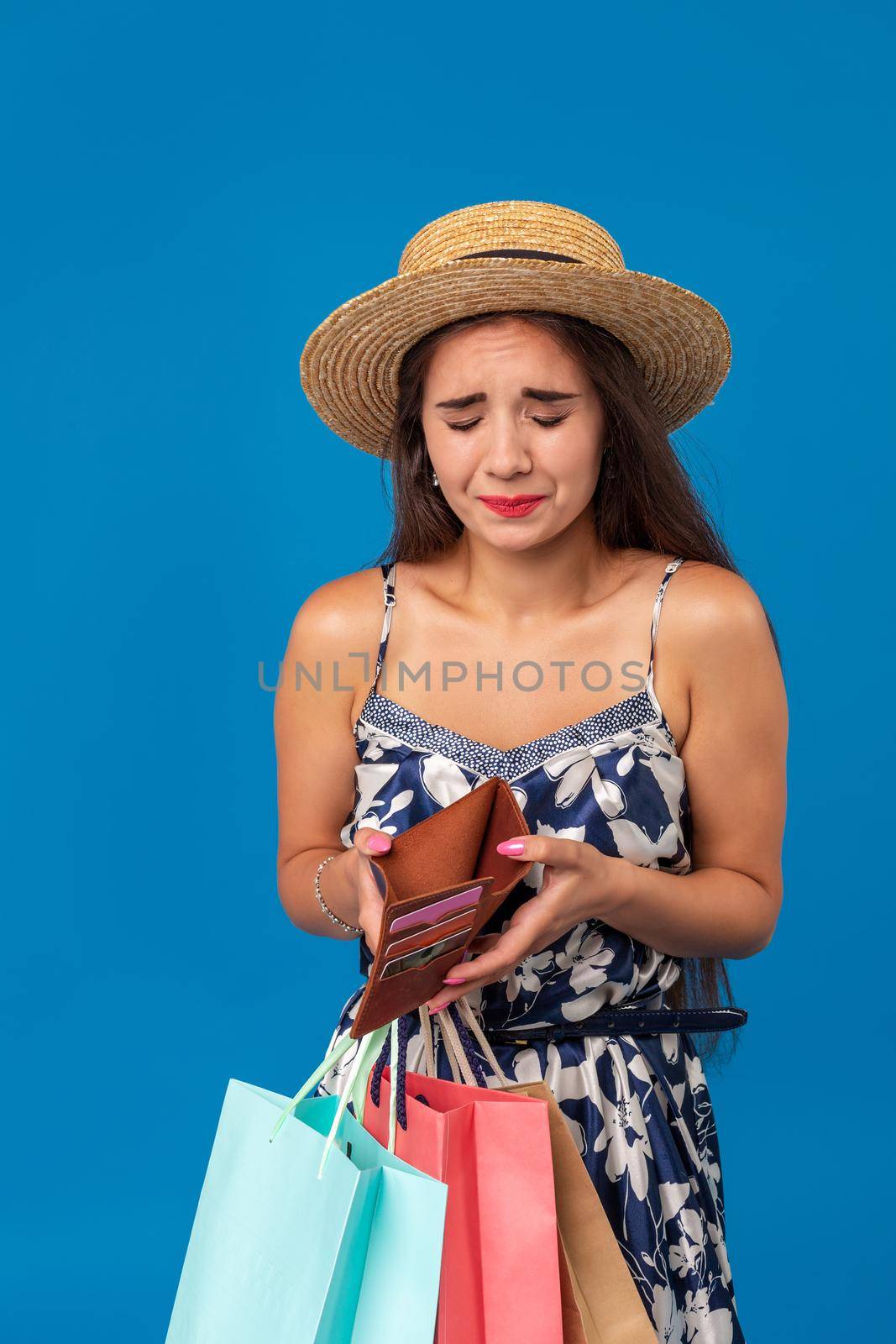 Portrait of unhappy young woman looking in her wallet in shopping center, spent too much, not enough cash, lost money, broke. Studio shot on blue background. Copy space