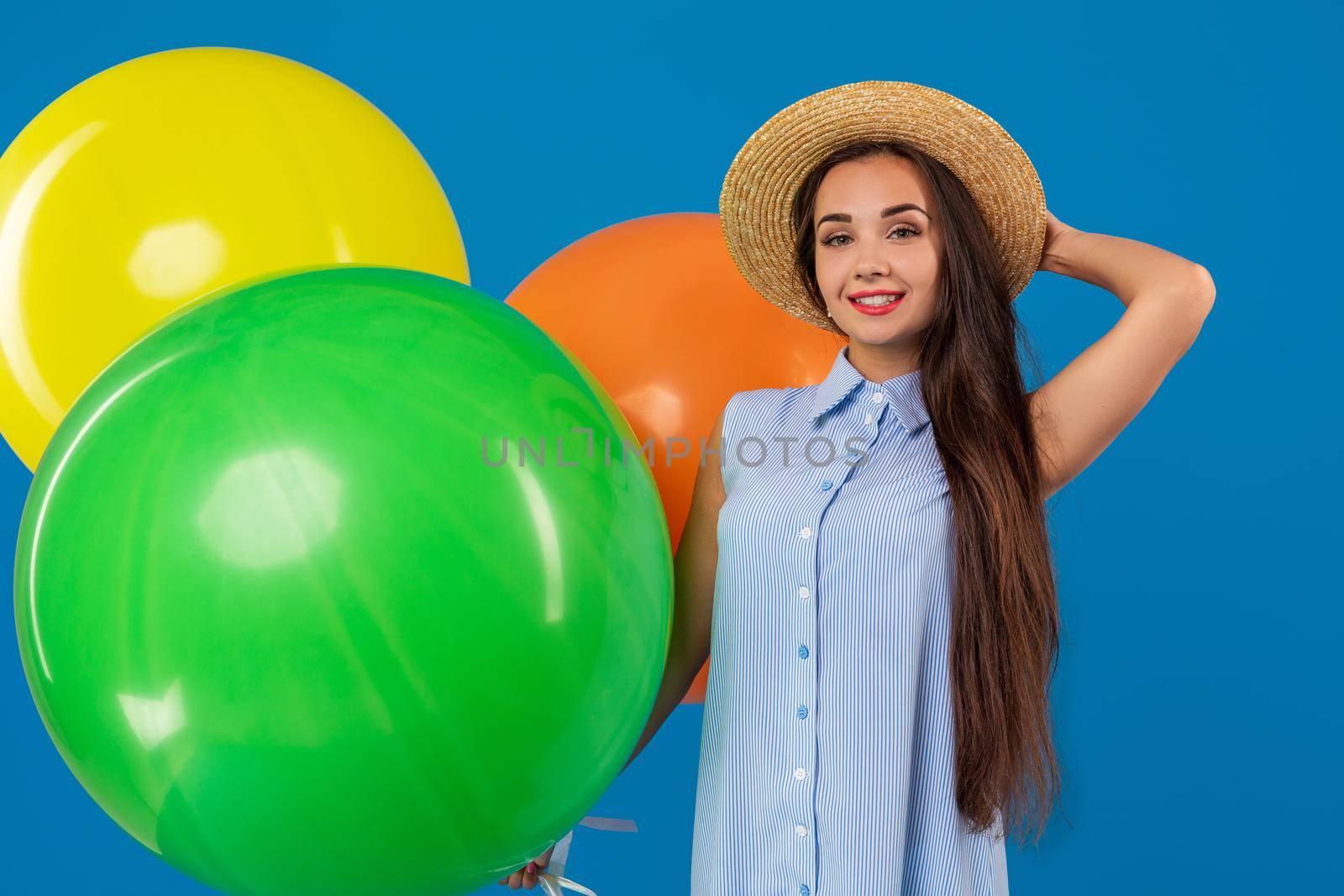 Cute brunette standing in a studio holding orange, yellow and green balloons. She wears light blue dress and has long and wavy hair. Celebration. Surprise