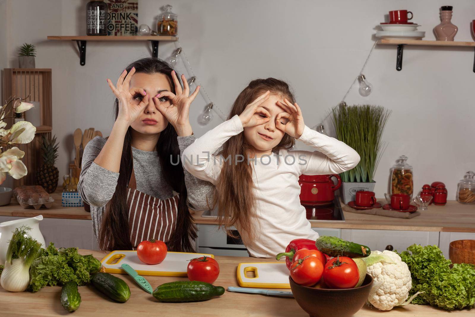 Happy loving family are cooking together. Loving mom and her kid are making a vegetable salad and fooling around at the kitchen, against a white wall with shelves and bulbs on it. Homemade food and little helper.